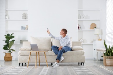 Photo of Man using remote to control air conditioner on sofa at home