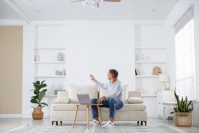 Photo of Man using remote to control air conditioner on sofa at home