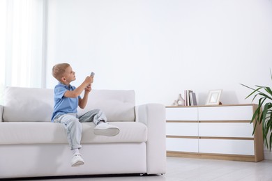 Photo of Cute little boy using remote to control air conditioner on sofa at home