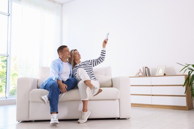 Photo of Man and woman using remote to control air conditioner on sofa at home