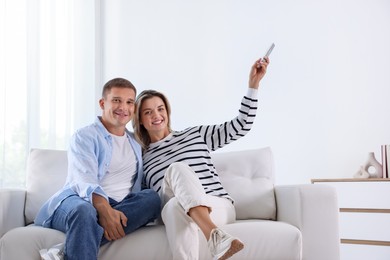 Photo of Man and woman using remote to control air conditioner on sofa at home