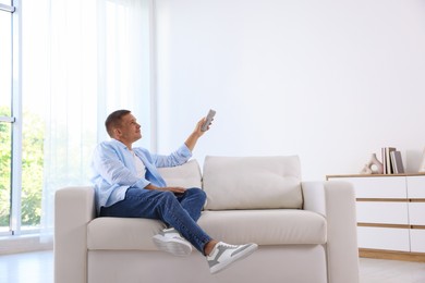 Photo of Man using remote to control air conditioner on sofa at home