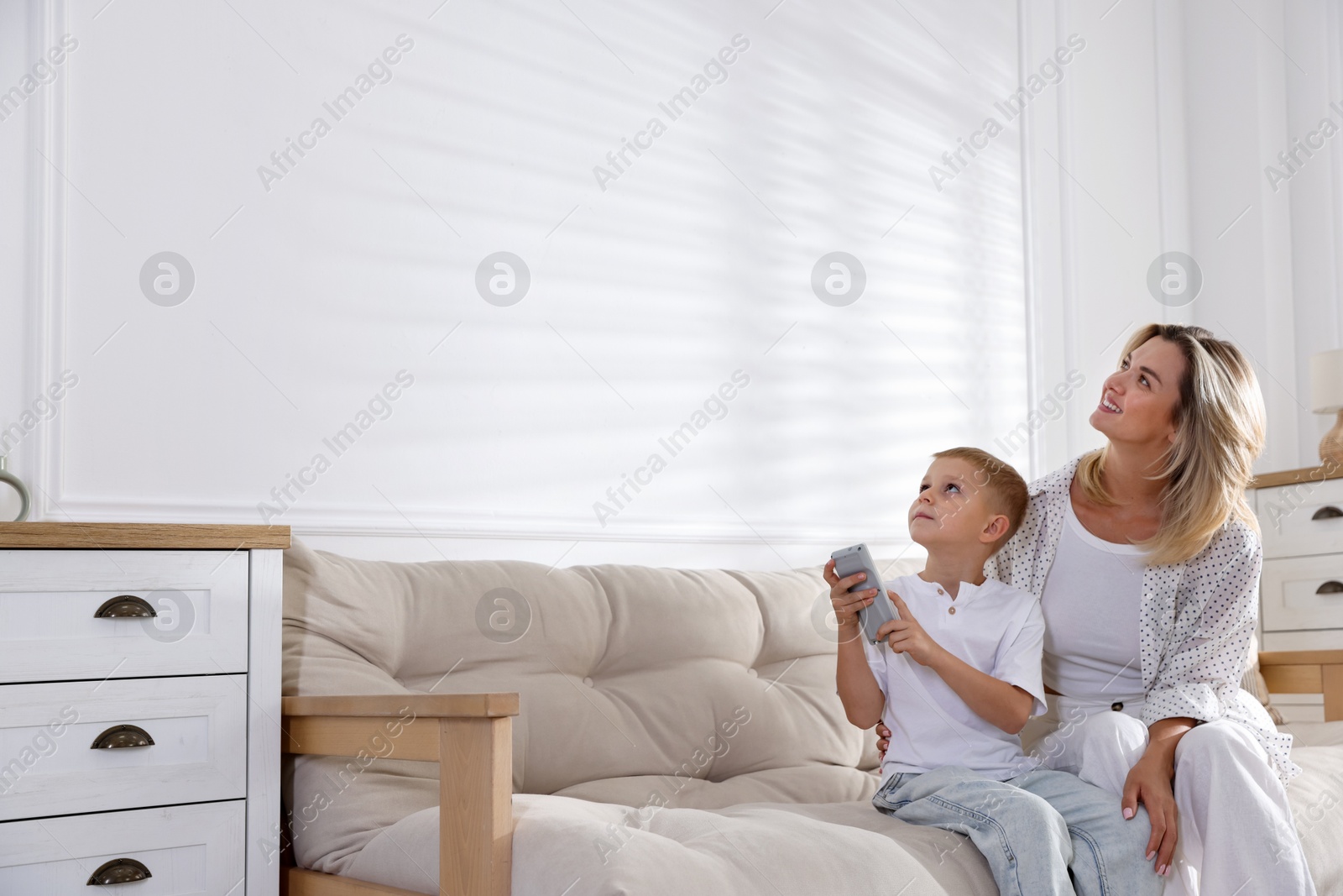 Photo of Woman and her son using remote to control air conditioner on sofa at home