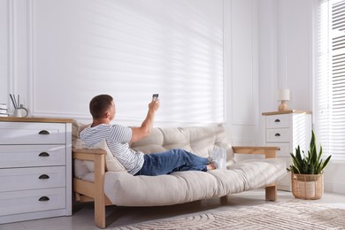 Photo of Man using remote to control air conditioner on sofa at home
