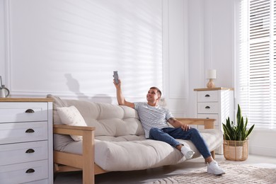 Photo of Man using remote to control air conditioner on sofa at home