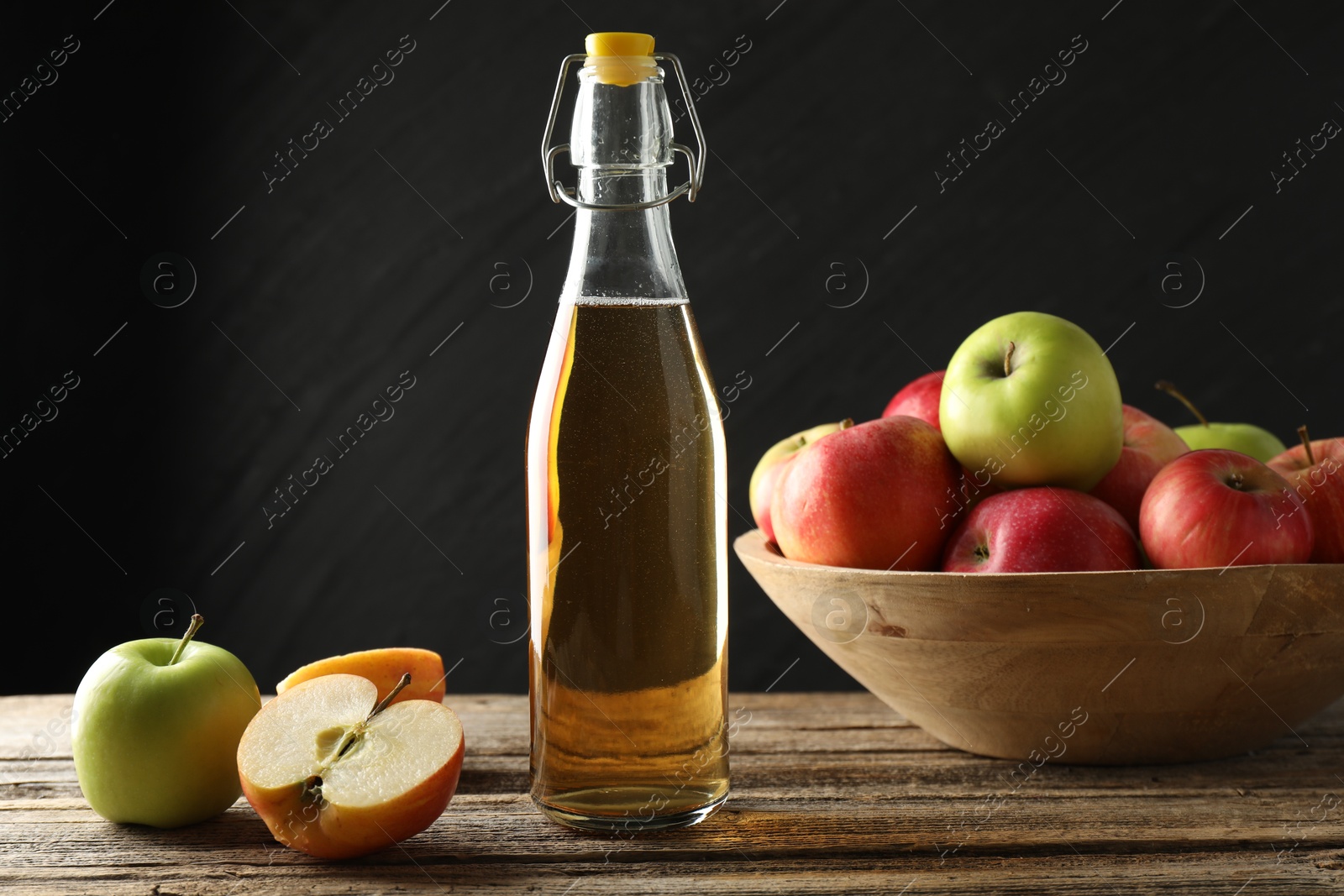 Photo of Delicious cider in glass bottle and apples on wooden table against dark gray background