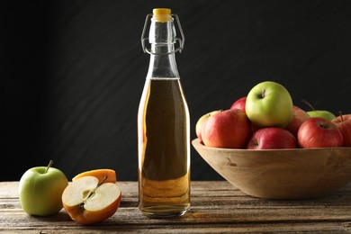 Photo of Delicious cider in glass bottle and apples on wooden table against dark gray background