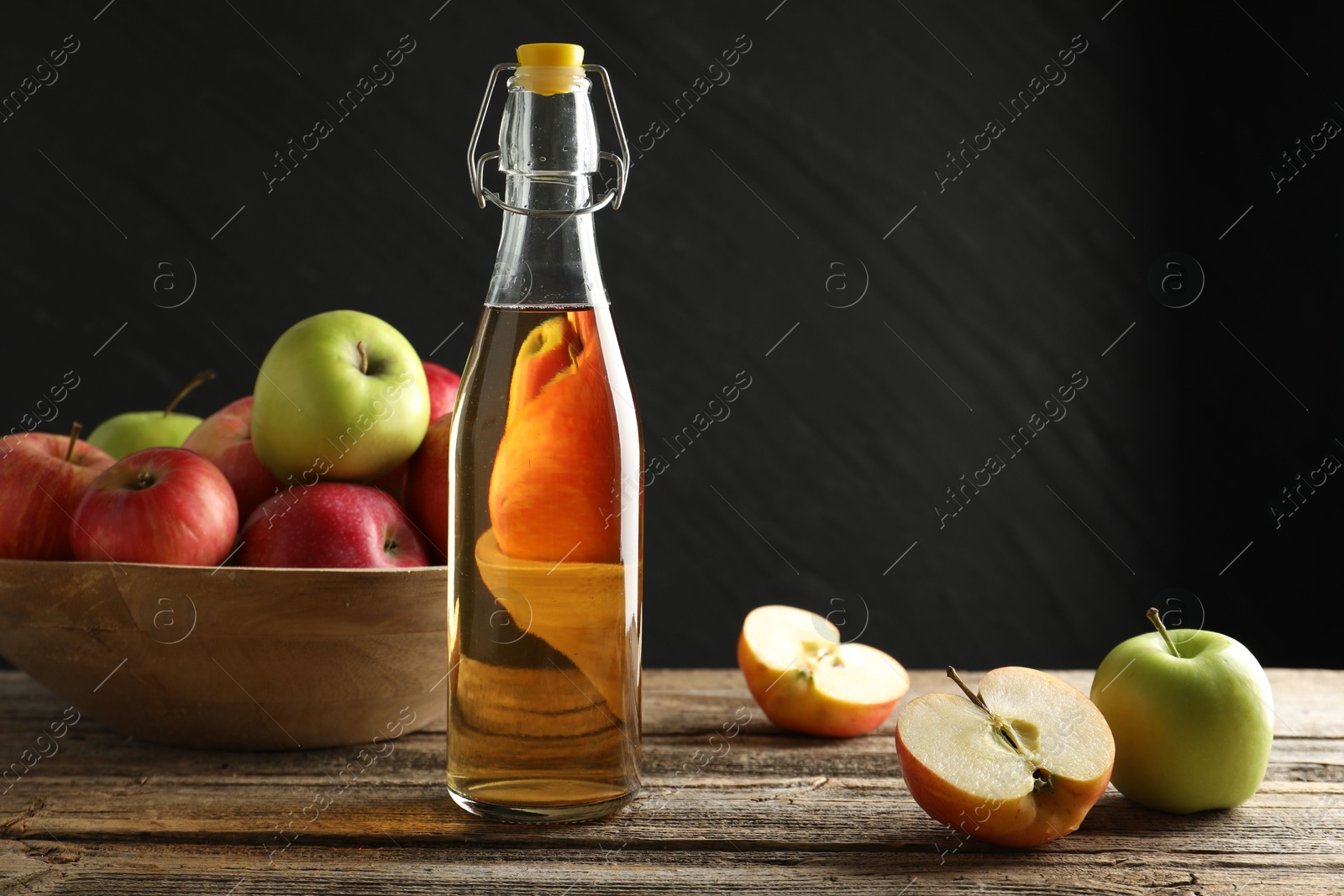 Photo of Delicious cider in glass bottle and apples on wooden table against dark gray background. Space for text
