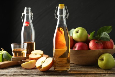 Photo of Delicious cider in glass bottles and apples on wooden table against dark gray background