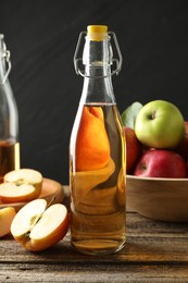 Photo of Delicious cider in glass bottle and apples on wooden table against dark gray background, closeup