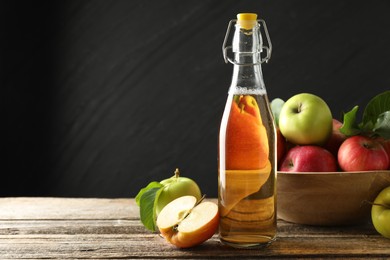 Photo of Delicious cider in glass bottle and apples on wooden table against dark gray background. Space for text
