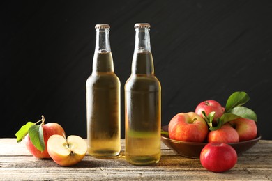 Photo of Delicious cider in glass bottles and apples on wooden table against dark gray background