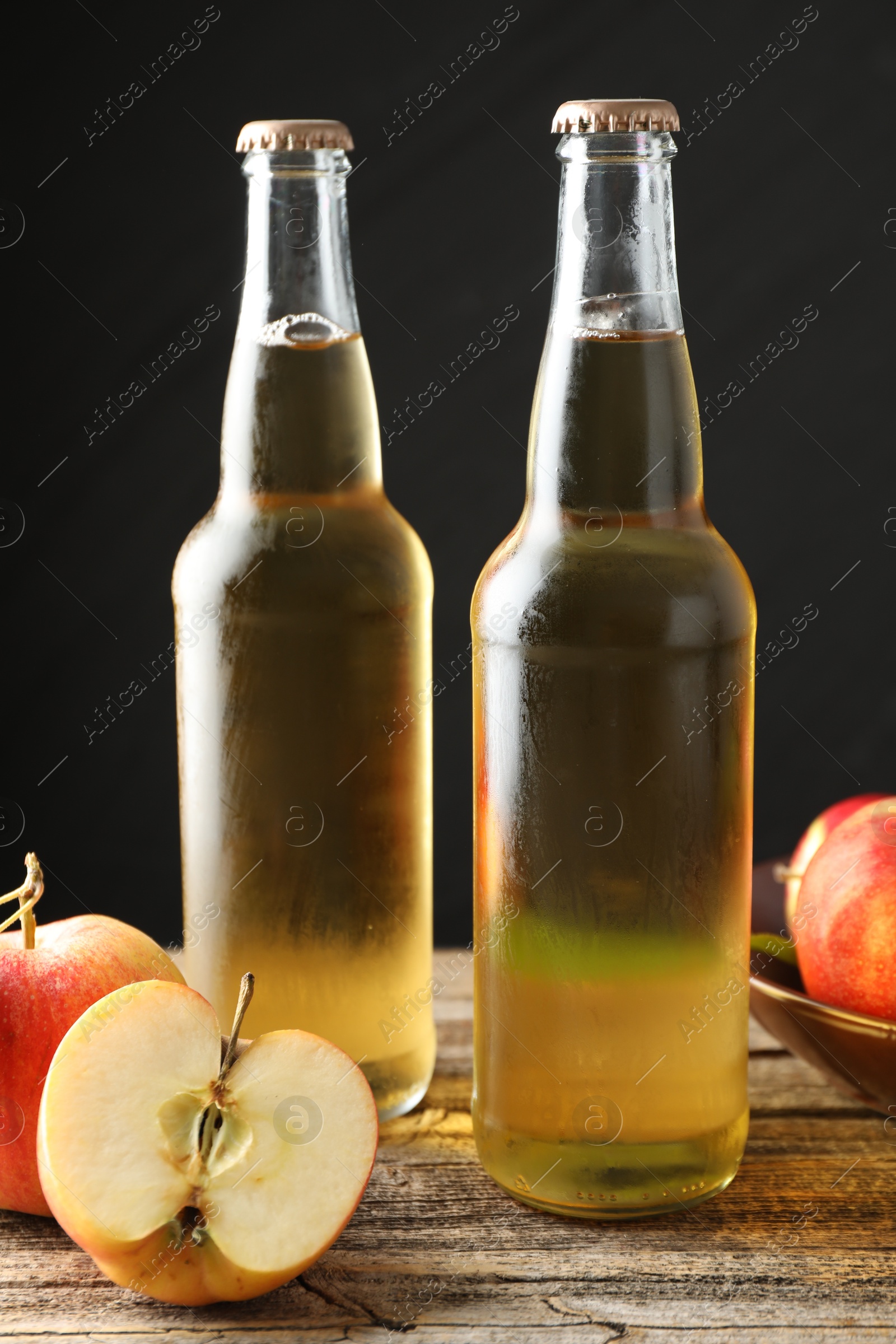 Photo of Delicious cider in glass bottles and apples on wooden table against dark gray background, closeup