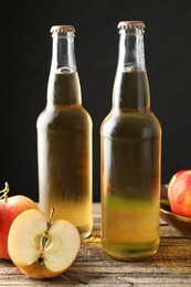 Photo of Delicious cider in glass bottles and apples on wooden table against dark gray background, closeup