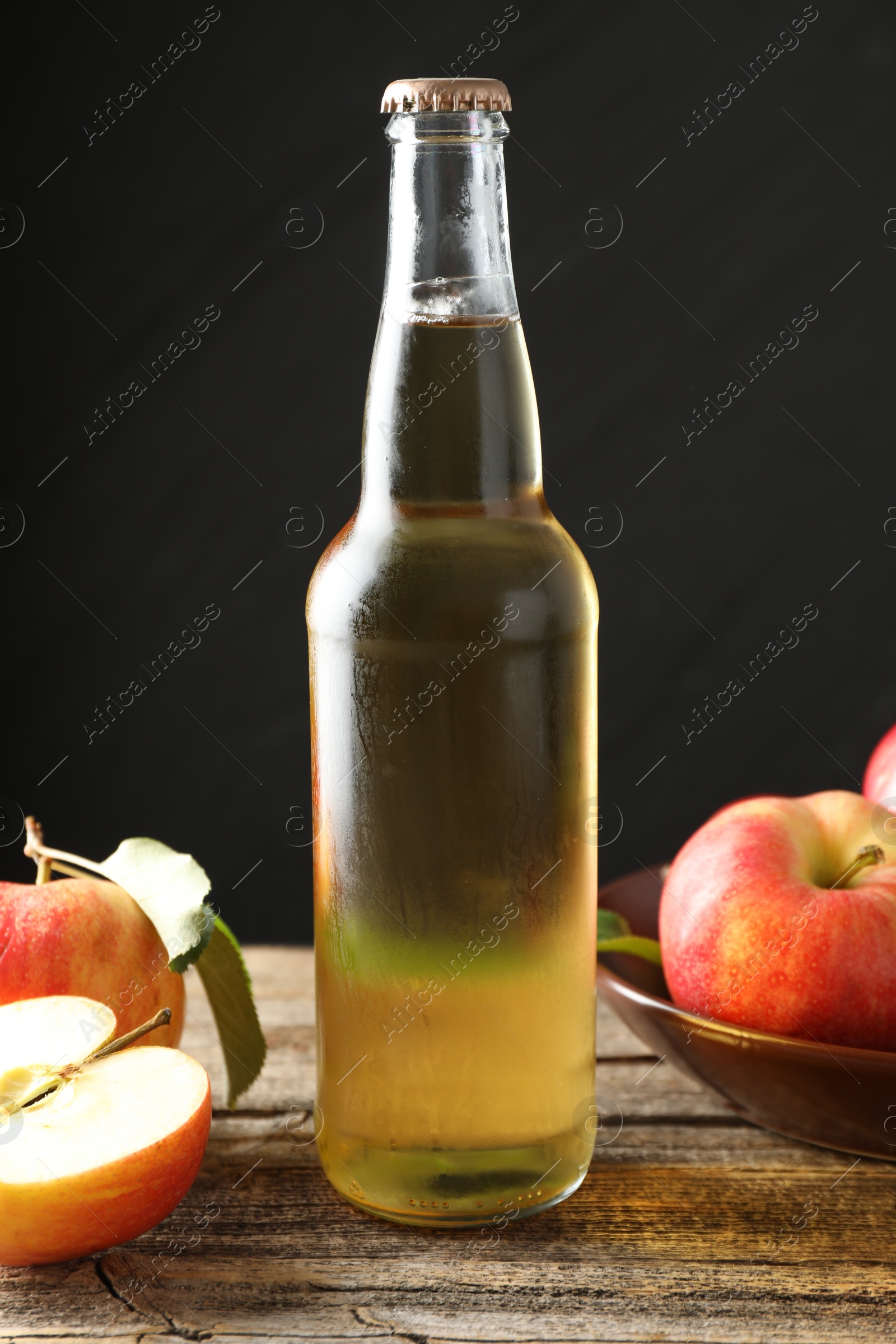 Photo of Delicious cider in glass bottle and apples on wooden table against dark gray background, closeup