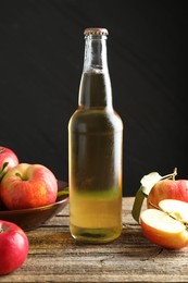 Photo of Delicious cider in glass bottle and apples on wooden table against dark gray background