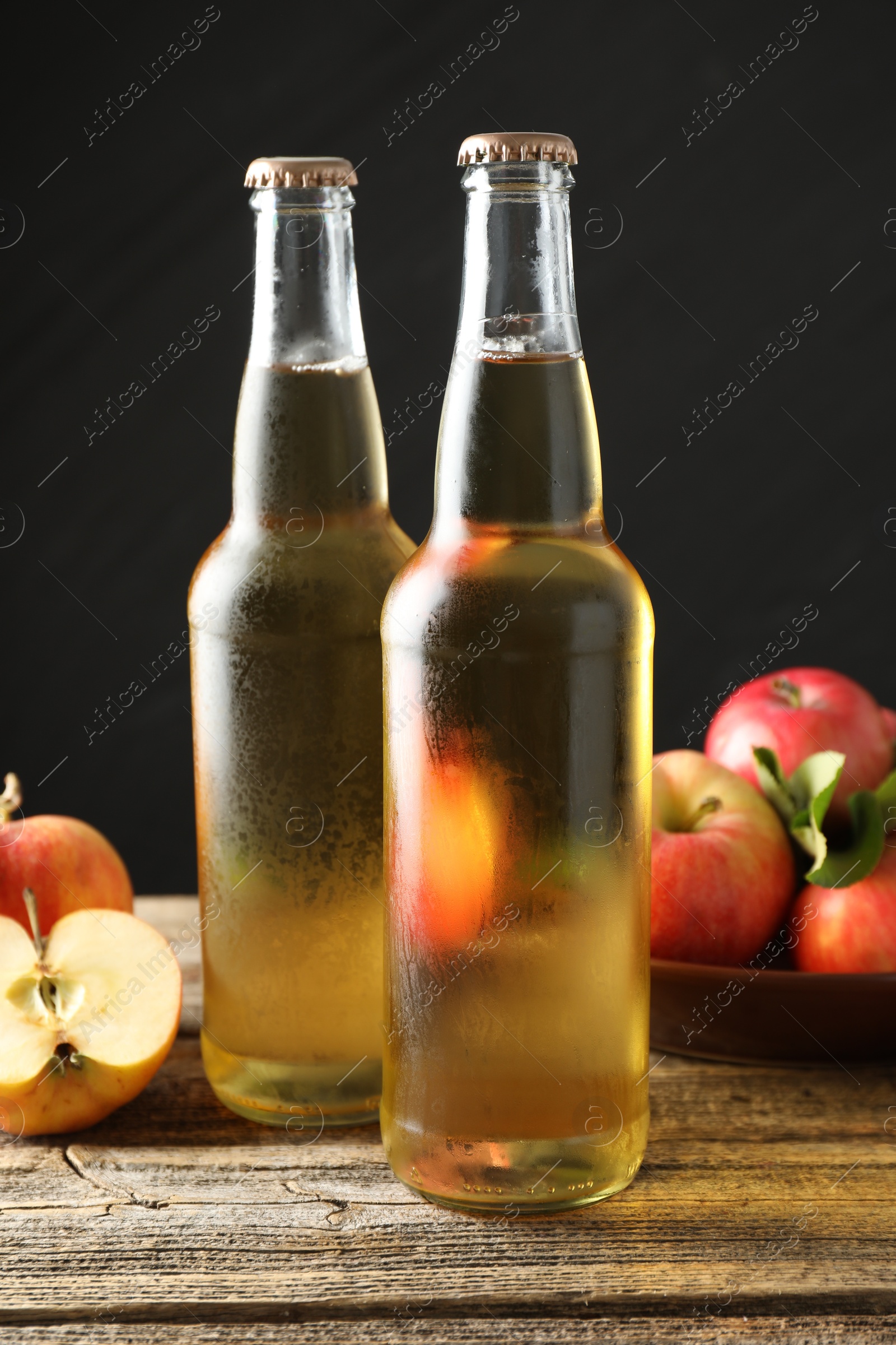 Photo of Delicious cider in glass bottles and apples on wooden table against dark gray background, closeup