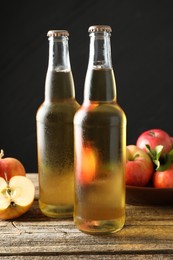 Photo of Delicious cider in glass bottles and apples on wooden table against dark gray background, closeup