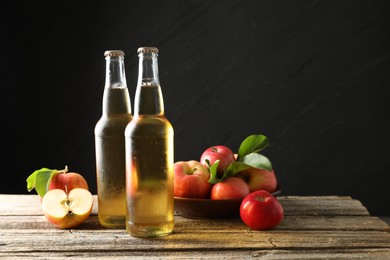 Photo of Delicious cider in glass bottles and apples on wooden table against dark gray background. Space for text