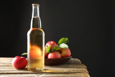 Photo of Delicious cider in glass bottle and apples on wooden table against dark gray background. Space for text