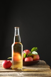 Photo of Delicious cider in glass bottle and apples on wooden table against dark gray background
