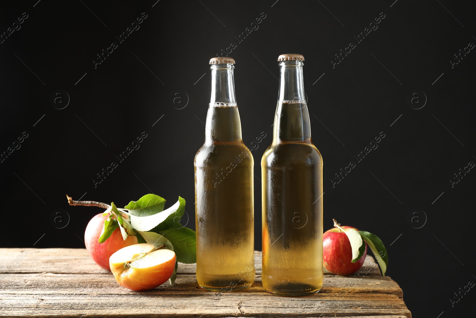 Photo of Delicious cider in glass bottles and apples on wooden table against dark gray background