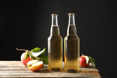Photo of Delicious cider in glass bottles and apples on wooden table against dark gray background