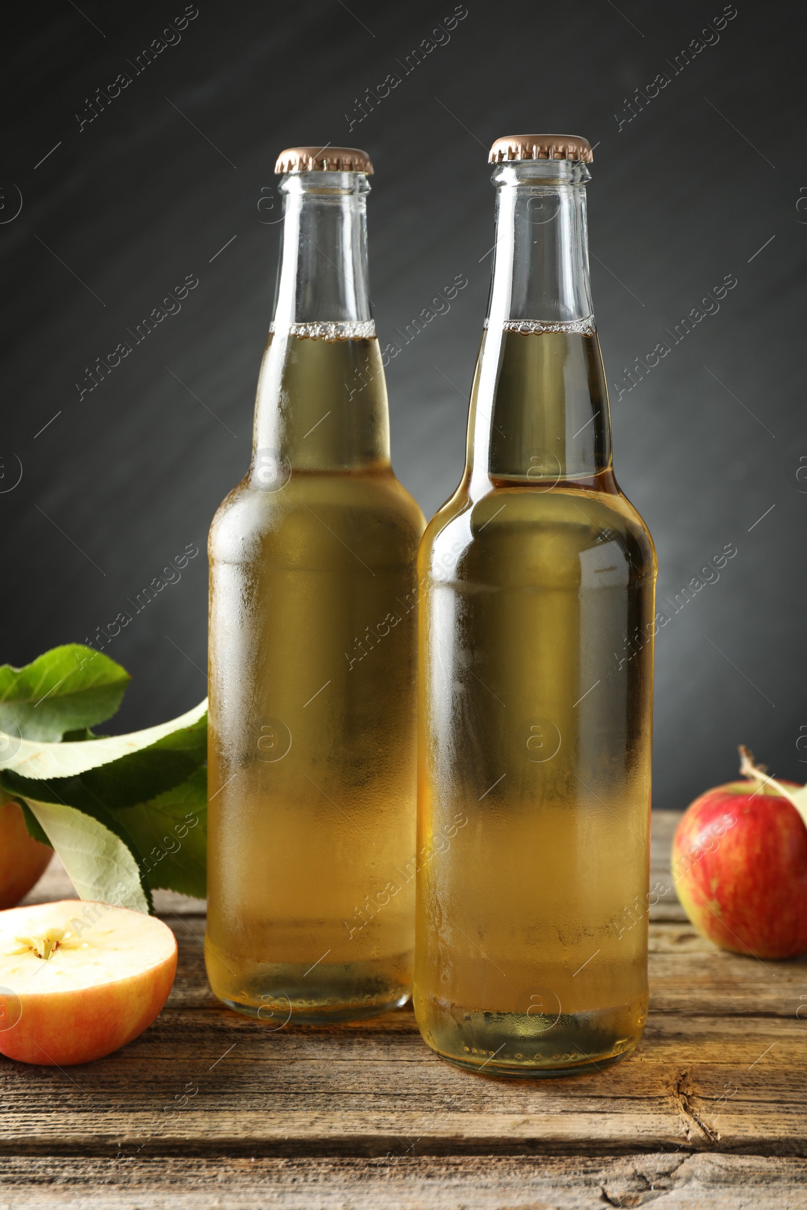 Photo of Delicious cider in glass bottles and apples on wooden table against dark gray background