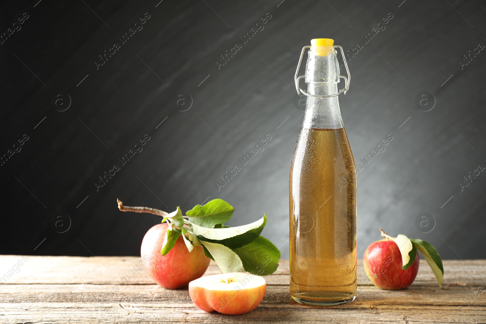 Photo of Delicious cider in glass bottle and apples on wooden table against dark gray background. Space for text