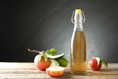 Photo of Delicious cider in glass bottle and apples on wooden table against dark gray background. Space for text
