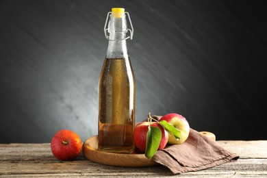 Photo of Delicious cider in glass bottle and apples on wooden table against dark gray background