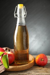 Photo of Delicious cider in glass bottle and apples on wooden table against dark gray background
