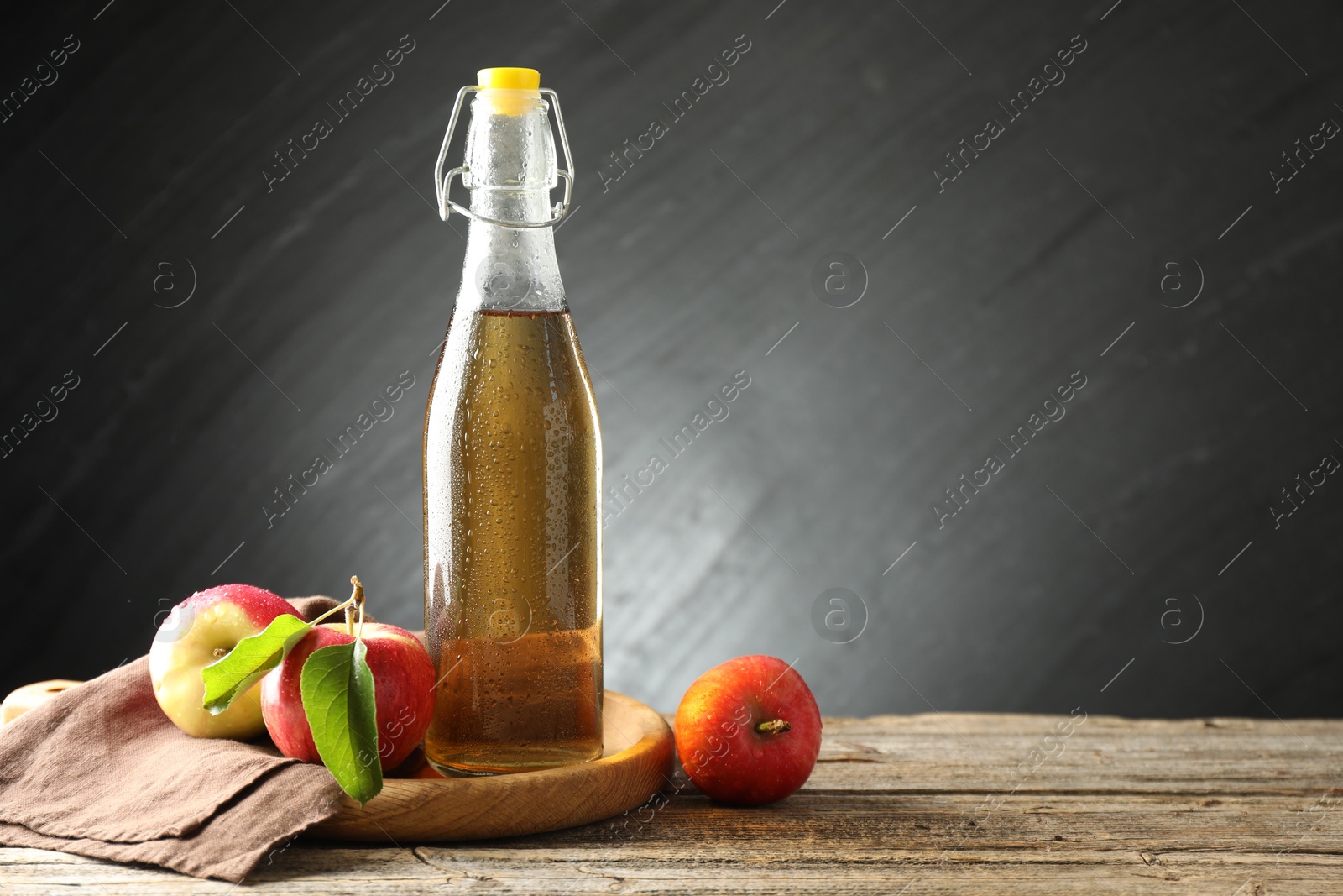 Photo of Delicious cider in glass bottle and apples on wooden table against dark gray background. Space for text