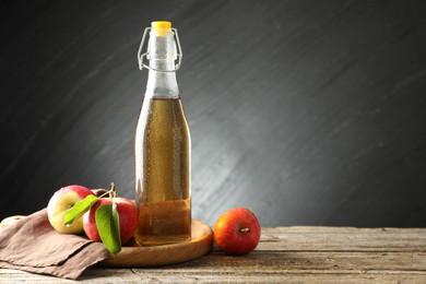 Photo of Delicious cider in glass bottle and apples on wooden table against dark gray background. Space for text