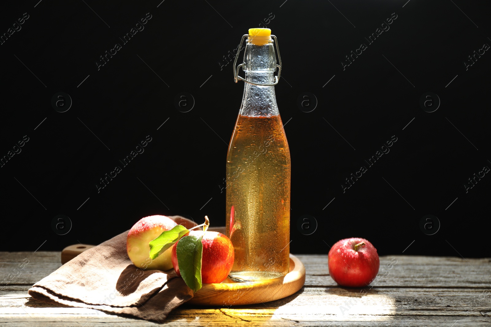 Photo of Delicious cider in glass bottle and apples on wooden table against dark background