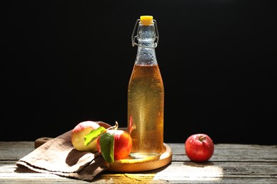 Photo of Delicious cider in glass bottle and apples on wooden table against dark background