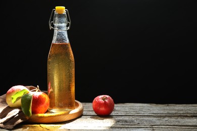 Photo of Delicious cider in glass bottle and apples on wooden table against dark background. Space for text