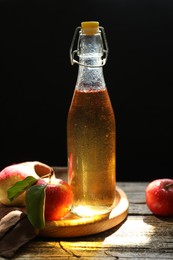 Photo of Delicious cider in glass bottle and apples on wooden table against dark background, closeup
