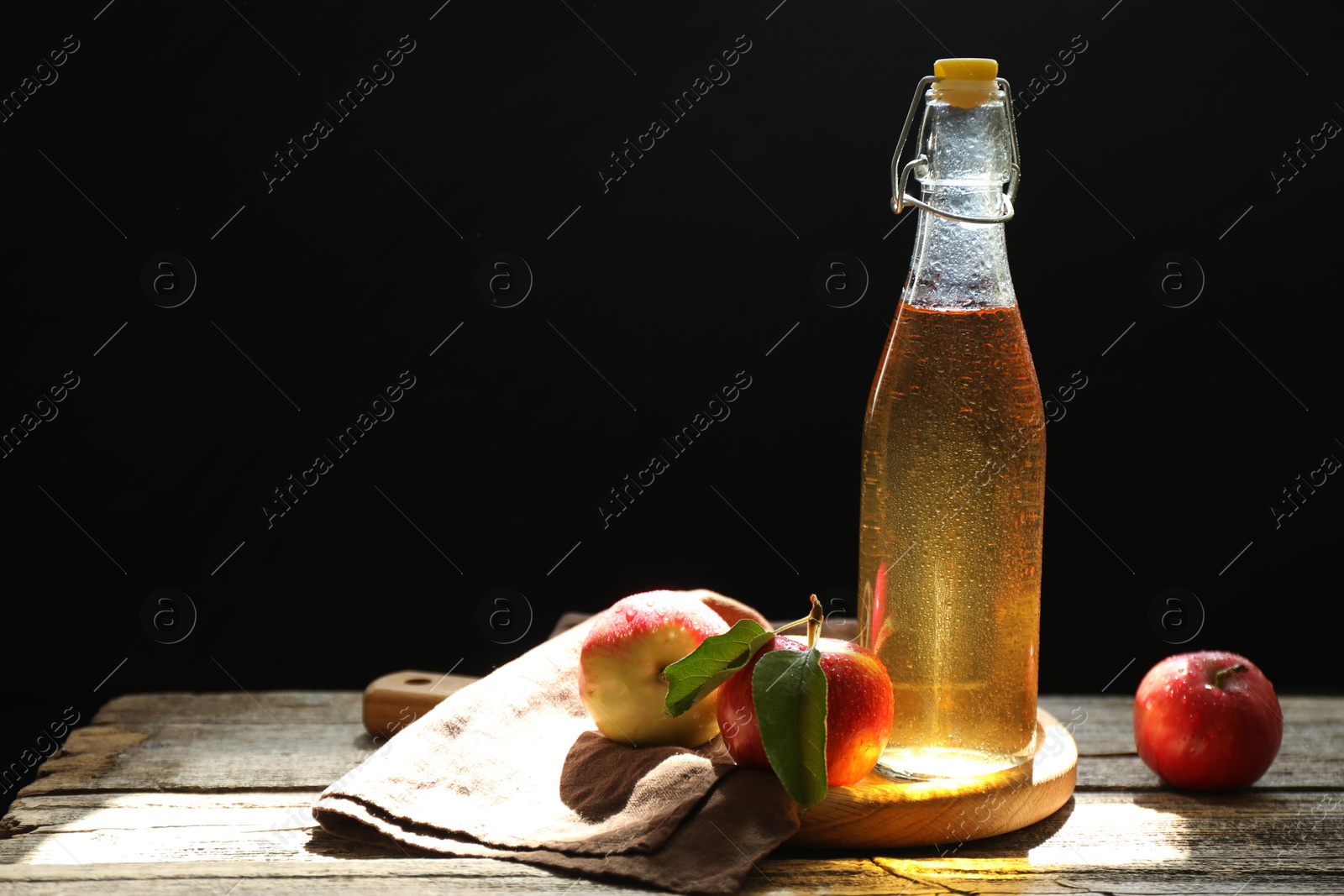 Photo of Delicious cider in glass bottle and apples on wooden table against dark background. Space for text