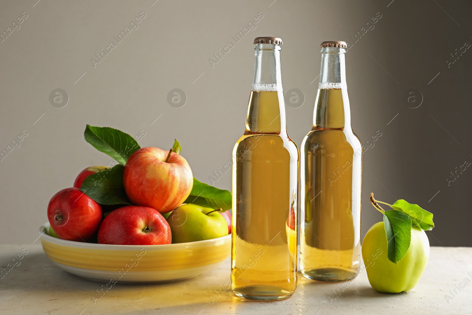 Photo of Delicious cider in glass bottles and apples on gray table against light background