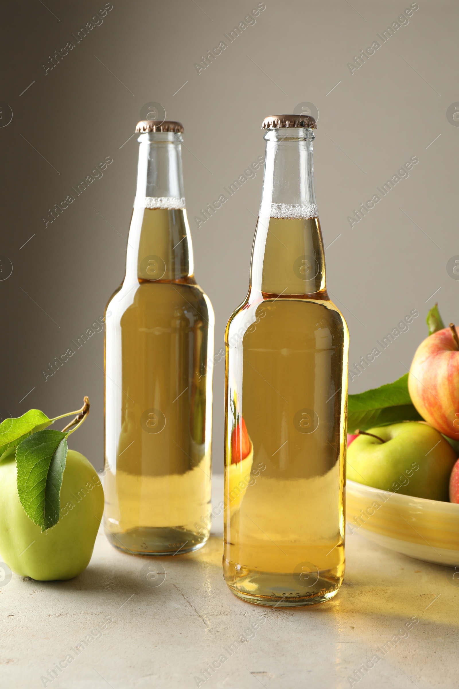 Photo of Delicious cider in glass bottles and apples on gray table against light background