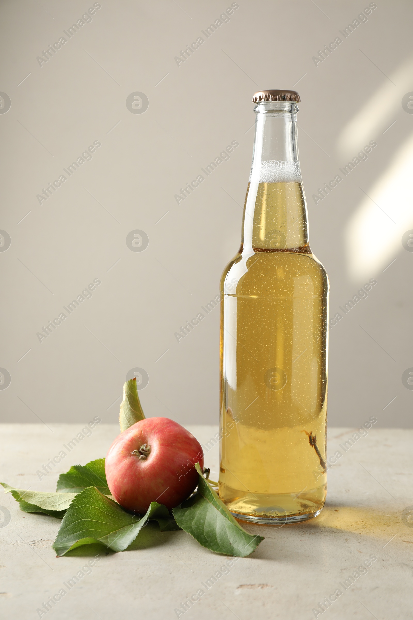 Photo of Delicious cider in glass bottle and apples on gray table against light background