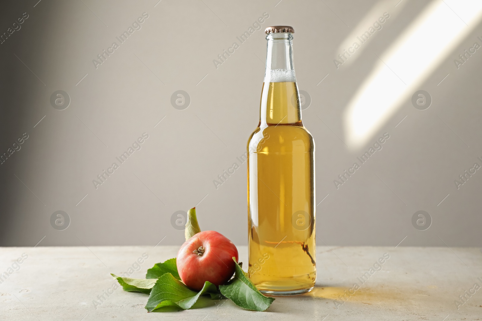 Photo of Delicious cider in glass bottle and apples on gray table against light background