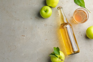 Photo of Delicious cider in bottle, glass and apples with green leaves on gray textured table, flat lay. Space for text