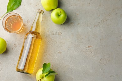 Photo of Delicious cider in bottle, glass and apples with green leaves on gray textured table, flat lay. Space for text