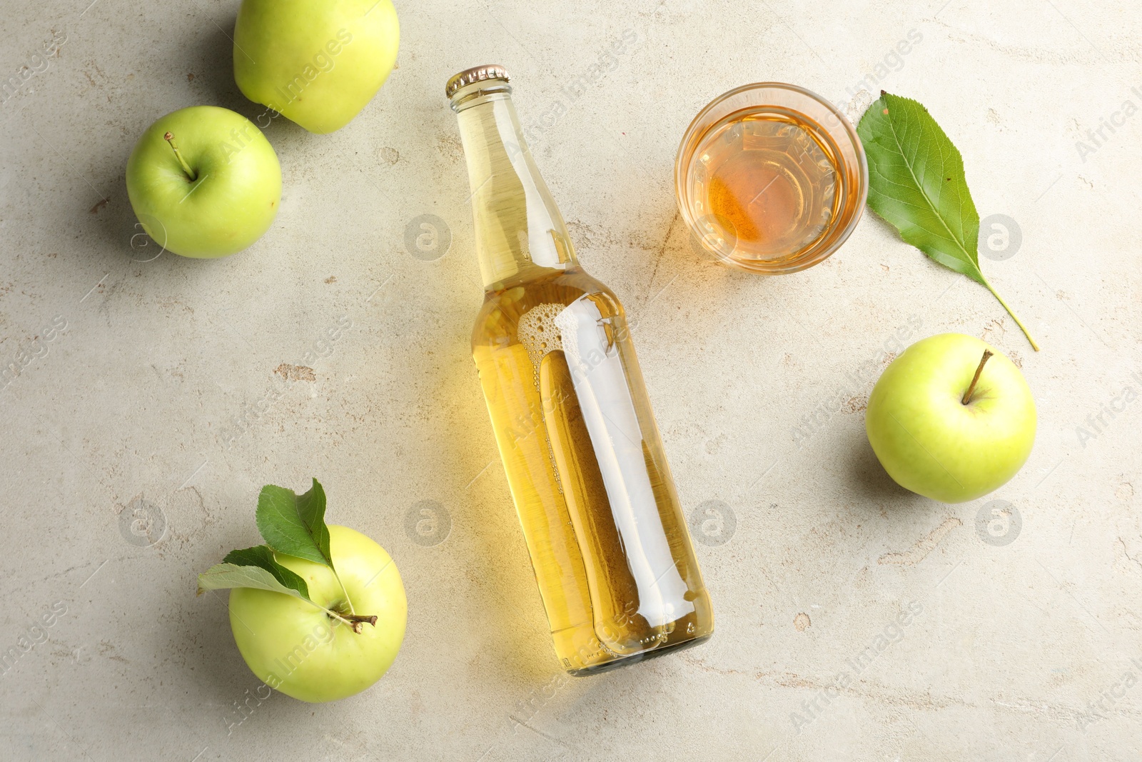 Photo of Delicious cider in bottle, glass and apples with green leaves on gray textured table, flat lay