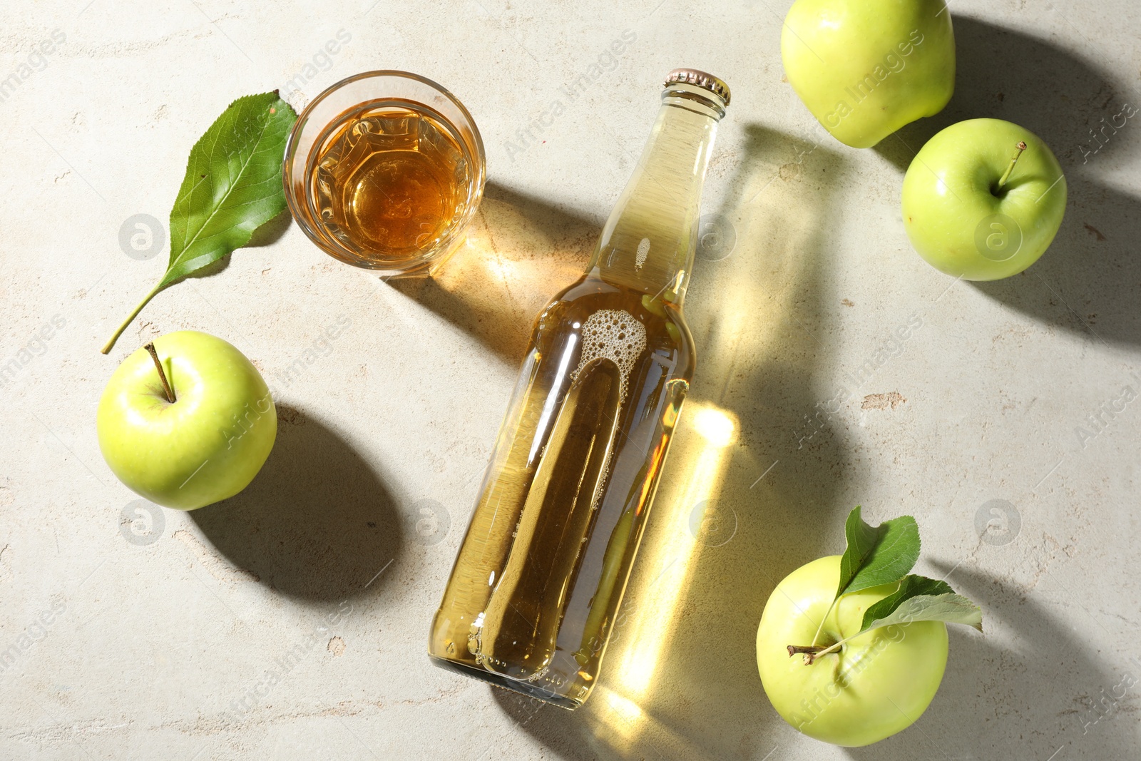 Photo of Delicious cider in bottle, glass and apples with green leaves on gray textured table, flat lay