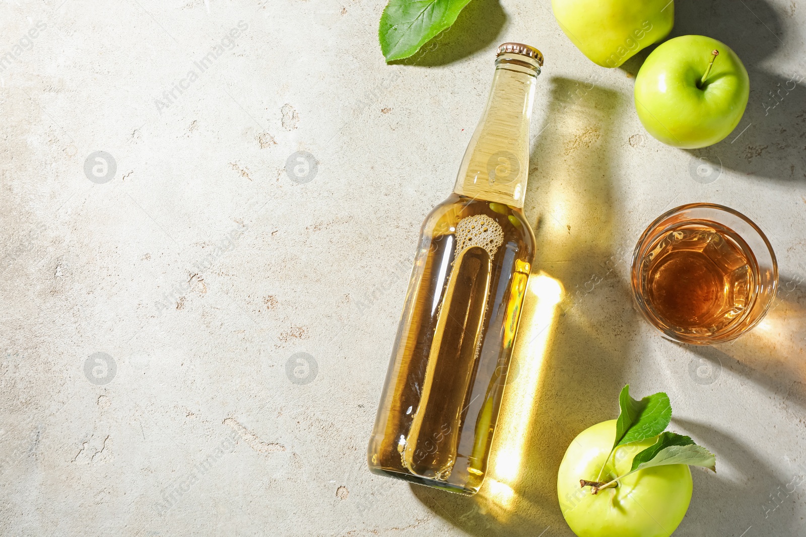 Photo of Delicious cider in bottle, glass and apples with green leaves on gray textured table, flat lay. Space for text