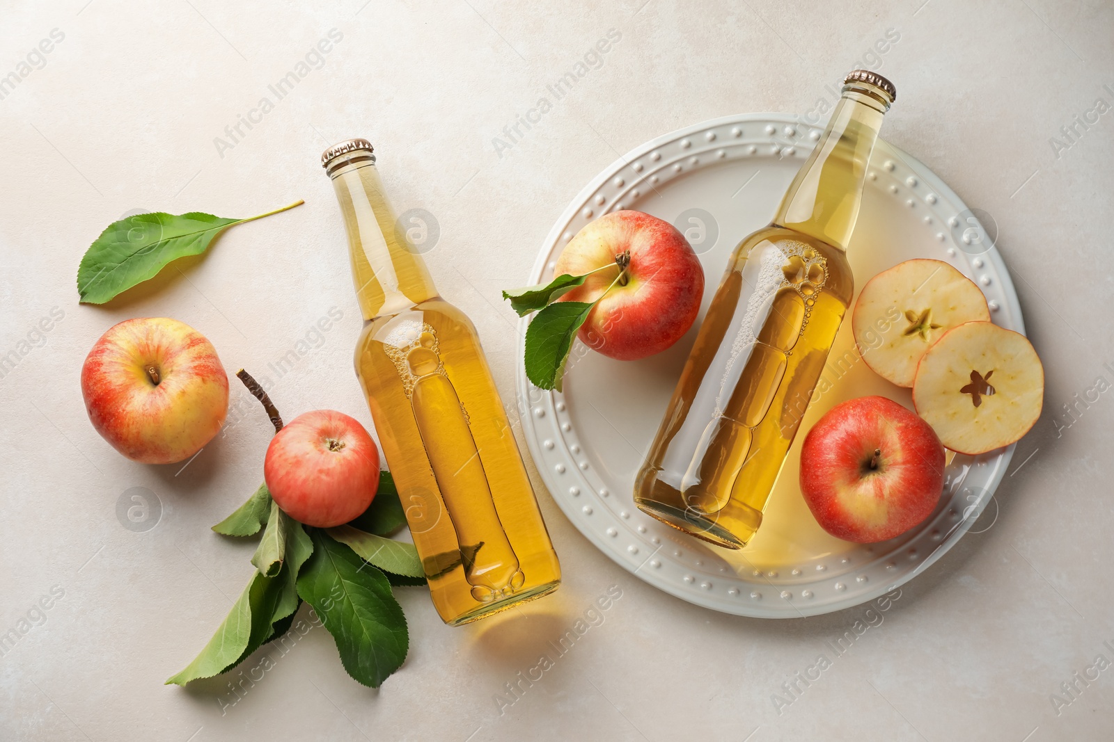 Photo of Delicious cider in glass bottles and apples with green leaves on beige table, flat lay