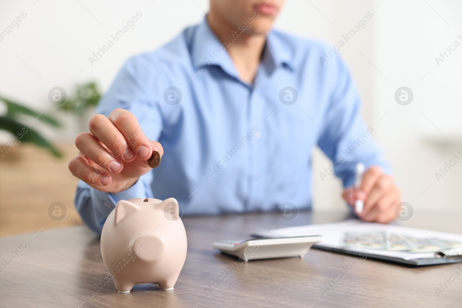 Photo of Man planning budget and putting money into piggy bank at wooden table, selective focus